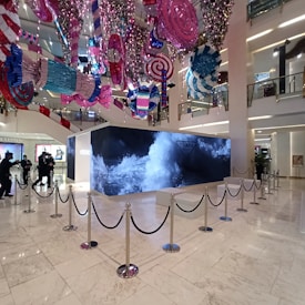 A retail store interior features a large Prada display surrounded by stanchions, with decorative, oversized candy-like ornaments hanging from the ceiling in vibrant colors. The floor is polished white marble, and the store has high ceilings with multiple floors visible. People are seen in the background near other luxury brand displays.