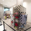 A retail display area featuring a collection of clothing items and accessories, including folded shirts, striped shirts on hangers, and various casual footwear. The display units are decorated with large black polka dots on a cream background, and there are sections labeled 'POCKET' with branding. The store has a modern and sleek design with concrete floors and bright lighting.
