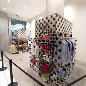 A retail display area featuring a collection of clothing items and accessories, including folded shirts, striped shirts on hangers, and various casual footwear. The display units are decorated with large black polka dots on a cream background, and there are sections labeled 'POCKET' with branding. The store has a modern and sleek design with concrete floors and bright lighting.