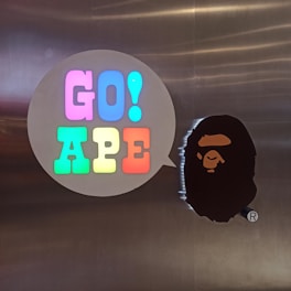 A metallic background features a speech bubble shape with brightly colored letters spelling 'GO! APE'. The letters are illuminated in pink, blue, green, yellow, and red. Next to the speech bubble is a silhouette of an ape's head in black.