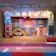 A colorful and vibrant stage design featuring geometric patterns and abstract shapes. The central figure has a stylized face with a hat, surrounded by various symmetrical designs in bright colors. The stage is lit with spotlights and decorated with a banner that reads 'EMJOY Empower the Youth's Nextperience'.