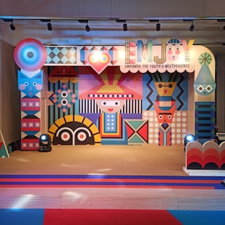 A colorful and vibrant stage design featuring geometric patterns and abstract shapes. The central figure has a stylized face with a hat, surrounded by various symmetrical designs in bright colors. The stage is lit with spotlights and decorated with a banner that reads 'EMJOY Empower the Youth's Nextperience'.