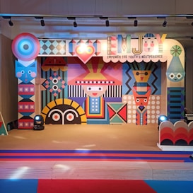 A colorful and vibrant stage design featuring geometric patterns and abstract shapes. The central figure has a stylized face with a hat, surrounded by various symmetrical designs in bright colors. The stage is lit with spotlights and decorated with a banner that reads 'EMJOY Empower the Youth's Nextperience'.
