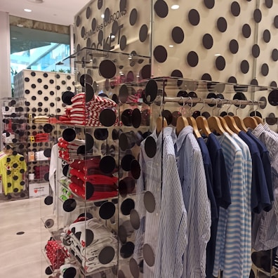 A clothing store display features a rack of neatly hung shirts in blue and white tones. Shelves nearby hold folded shirts, primarily in red and white patterns. The walls and display are decorated with large black polka dots, creating a bold and modern aesthetic. Visible through a glass pane, the layout is organized and stylish.