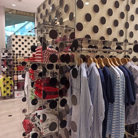 A clothing store display features a rack of neatly hung shirts in blue and white tones. Shelves nearby hold folded shirts, primarily in red and white patterns. The walls and display are decorated with large black polka dots, creating a bold and modern aesthetic. Visible through a glass pane, the layout is organized and stylish.
