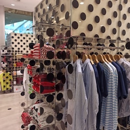 A clothing store display features a rack of neatly hung shirts in blue and white tones. Shelves nearby hold folded shirts, primarily in red and white patterns. The walls and display are decorated with large black polka dots, creating a bold and modern aesthetic. Visible through a glass pane, the layout is organized and stylish.