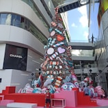 A large, festive holiday tree decorated with oversized candy-themed ornaments, including candy canes, lollipops, and colorful circular designs. The tree is set in a modern urban environment with adjacent glass buildings and branded storefronts. A child stands in front of the tree, adding to the lively atmosphere.