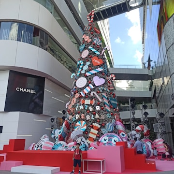 A large, festive holiday tree decorated with oversized candy-themed ornaments, including candy canes, lollipops, and colorful circular designs. The tree is set in a modern urban environment with adjacent glass buildings and branded storefronts. A child stands in front of the tree, adding to the lively atmosphere.