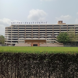 A multi-story building with a cream and black color scheme is visible. The structure features a sign with text written in a script that appears to be Thai, prominently displayed on the roof. In front of the building, there is an open grassy field separated by a trimmed green hedge. Several trees are positioned to the right of the building, adding some greenery to the scene. The sky is partly cloudy.