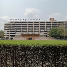 A multi-story building with a cream and black color scheme is visible. The structure features a sign with text written in a script that appears to be Thai, prominently displayed on the roof. In front of the building, there is an open grassy field separated by a trimmed green hedge. Several trees are positioned to the right of the building, adding some greenery to the scene. The sky is partly cloudy.
