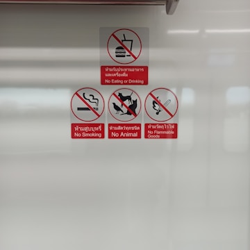 A set of four warning signs displayed on a flat surface. The signs include no eating or drinking, no smoking, no animals, and no flammable goods. Each is represented with a relevant icon and a red prohibition symbol, accompanied by text in both English and Thai.
