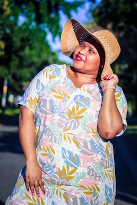 A person wearing a colorful dress with leaf patterns and a wide-brimmed straw hat poses outdoors, smiling in the sunlight. The background features blurred greenery, suggesting a park or garden setting.