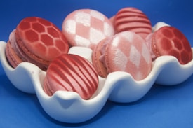 A set of beautifully decorated macarons with various pink patterns, including stripes and diamond shapes, arranged in a white, wavy dish against a blue background.