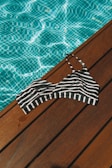 A black and white striped bikini top lies on a wooden deck beside a pool filled with clear blue water. The sunlight casts a shimmering pattern on the pool's surface, highlighting the contrast between the wood, the stripes, and the water.