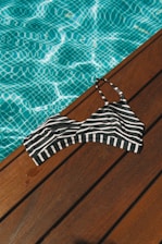 A black and white striped bikini top lies on a wooden deck beside a pool filled with clear blue water. The sunlight casts a shimmering pattern on the pool's surface, highlighting the contrast between the wood, the stripes, and the water.
