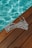 A black and white striped bikini top lies on a wooden deck beside a pool filled with clear blue water. The sunlight casts a shimmering pattern on the pool's surface, highlighting the contrast between the wood, the stripes, and the water.