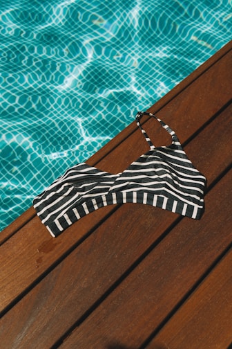 A black and white striped bikini top lies on a wooden deck beside a pool filled with clear blue water. The sunlight casts a shimmering pattern on the pool's surface, highlighting the contrast between the wood, the stripes, and the water.