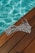 A black and white striped bikini top lies on a wooden deck beside a pool filled with clear blue water. The sunlight casts a shimmering pattern on the pool's surface, highlighting the contrast between the wood, the stripes, and the water.