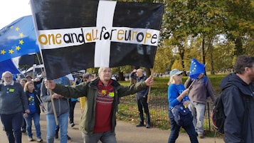 A group of people are participating in a public march or protest, walking along a path lined with trees. One person is holding a large black flag with a white cross and the words 'one and all for Europe' written in bold letters. Other individuals are carrying EU flags with yellow stars on a blue background. The participants are dressed in casual attire, some with badges or stickers indicating their support for Europe.