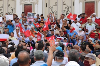 A large group of people gathered in a public space holding red flags with a white symbol, likely depicting a political or social demonstration. The crowd is diverse, with some individuals wearing masks and others holding signs and banners. Many people are taking photos or videos with their phones.