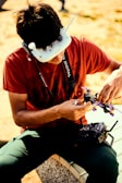 A person is seated outdoors wearing a white headset with antennas, likely for controlling a drone. They are holding a small drone or similar device in their hands and have a camera strap around their neck. The person is dressed in a red shirt and green pants, and there is a remote control device on their lap.