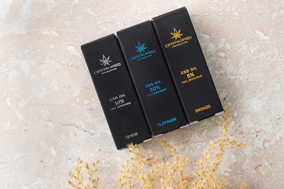 Three rectangular black boxes labeled with 'Crystalweed' are arranged on a textured beige surface. Each box displays different CBD oil concentrations: 10%, 30%, and 5%, with respective labels 'Silver', 'Platinum', and 'Bronze'. The boxes have subtle decorative elements resembling a cannabis leaf in different colors.