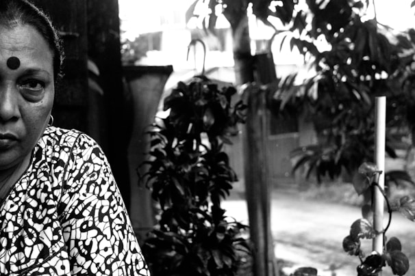 A woman with a patterned outfit and a bindi on her forehead is partially visible. She has a serious expression on her face. The background features foliage, including a plant with entwined vines around a pole, and a blurred view of a street.