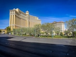 A large, luxurious hotel with classical architectural elements stands prominently against a clear blue sky. The building features a series of columns and ornate details in a cream color. In the foreground, a wide road bordered by well-maintained landscaping with shrubs and trees is visible.