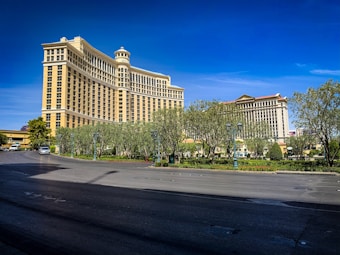 A large, luxurious hotel with classical architectural elements stands prominently against a clear blue sky. The building features a series of columns and ornate details in a cream color. In the foreground, a wide road bordered by well-maintained landscaping with shrubs and trees is visible.