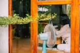 A woman is sitting indoors near a wooden-framed glass door, holding a phone. A fan is placed in front of her. Outside, greenery can be seen.