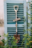 A decorative shovel with flowers and a 'Garden' sign hanging against a rustic, light blue shutter. Surrounding greenery adds a touch of nature to the scene.