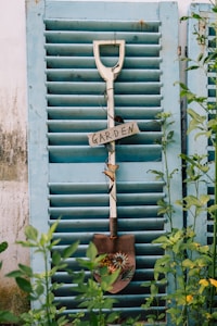 A decorative shovel with flowers and a 'Garden' sign hanging against a rustic, light blue shutter. Surrounding greenery adds a touch of nature to the scene.