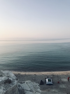 A tranquil beach scene with calm sea waters stretching to the horizon under a clear sky. The foreground features a rocky cliff overlooking a sandy shoreline. Two tents and a few camping chairs are set up near the water's edge, indicating a campsite. A person wearing a red shirt stands nearby, adding a subtle human presence to the natural landscape.