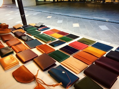 A display of various leather wallets arranged on a table, featuring different colors and styles. The table is situated outdoors with a tiled ground in the background.