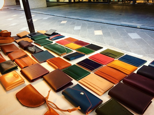 A display of various leather wallets arranged on a table, featuring different colors and styles. The table is situated outdoors with a tiled ground in the background.