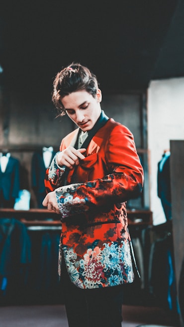 A person wearing an ornate, floral-patterned red jacket is adjusting the pocket square while standing in a clothing store. Various suits hang on racks in the background, creating a sophisticated atmosphere.