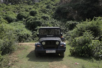 A black Mahindra jeep is parked on a grassy path surrounded by dense greenery and vegetation. The jeep is centered in the image, facing forward, with its headlights and distinctive grille visible. The background features a lush, wooded hillside, creating a natural and rugged setting.