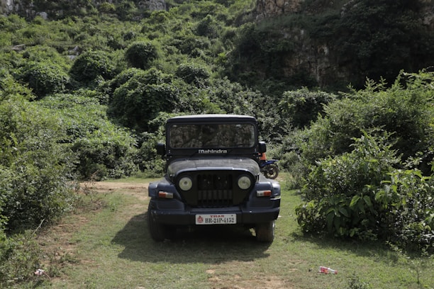 A black Mahindra jeep is parked on a grassy path surrounded by dense greenery and vegetation. The jeep is centered in the image, facing forward, with its headlights and distinctive grille visible. The background features a lush, wooded hillside, creating a natural and rugged setting.