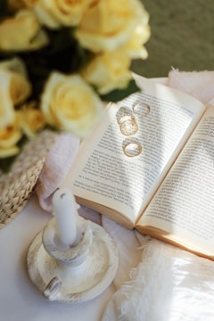 An open book lies on a table, surrounded by a woven basket filled with yellow roses. A white candle in a rustic holder rests nearby, and two wedding rings cast shadows on the book's pages. Sunlight softly illuminates the scene, creating a warm and intimate atmosphere.