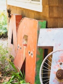 Large, weathered metal letters leaning against a wooden building, partially surrounded by greenery. A rusty bicycle wheel is positioned in front of the letters, and the scene conveys an outdoor, rustic setting with the building featuring brown wooden shingles.
