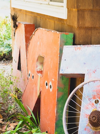 Large, weathered metal letters leaning against a wooden building, partially surrounded by greenery. A rusty bicycle wheel is positioned in front of the letters, and the scene conveys an outdoor, rustic setting with the building featuring brown wooden shingles.