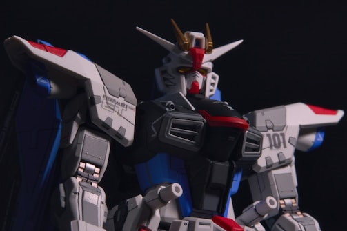 A detailed model of a mech robot with complex designs, featuring sharp angles and contrasting colors. The robot has a helmet-like head with a red face and yellow visor, and prominent horns protruding from the top. Its armor consists of white, gray, blue, and red sections, with intricate technical details and panels visible. The lighting casts strong shadows, emphasizing the mechanical structure.