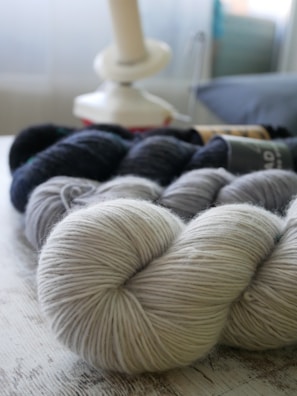A collection of yarn skeins in various shades of gray and black is arranged on a wooden surface. In the background, a yarn winder is visible, softly out of focus, contributing to a cozy and craft-oriented setting.