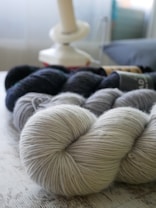 A collection of yarn skeins in various shades of gray and black is arranged on a wooden surface. In the background, a yarn winder is visible, softly out of focus, contributing to a cozy and craft-oriented setting.