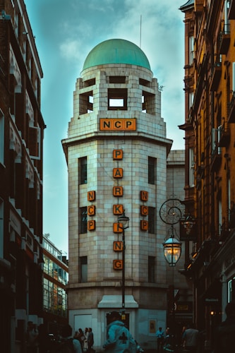 A tall, cylindrical building with a large sign that spells out 'NCP PARKING' using illuminated letters. The building is white with a green dome at the top and is situated between two other structures. Street lamps with intricate designs can be seen on the sides and several pedestrians are present near the entrance.