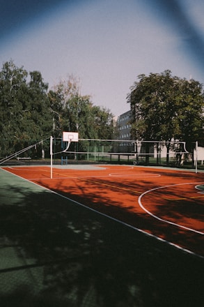 A basketball court with a red playing surface, located outdoors and surrounded by trees. The court has a basketball hoop and lines painted for various sports. The area is shadowed by trees, and a building is visible in the background.