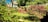 A lush garden with a variety of greenery, including a small red shed with white-framed windows. The garden features neatly trimmed grass, various bushes, and small trees. There is a decorative metal structure and a background with a white multi-story house and additional trees.