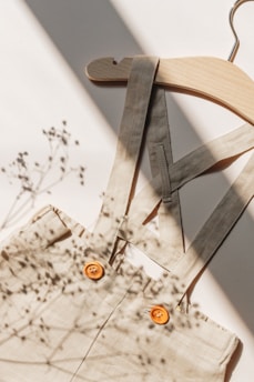 A wooden hanger holds a beige garment with two orange buttons. The fabric of the garment features shadows of delicate plants, suggesting a serene and natural setting. The light source from the side creates soft, contrasting shadows.