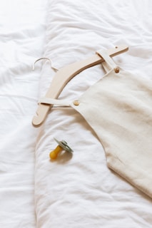 A wooden hanger holds a beige linen garment, laid out on a soft white surface, next to a yellow pacifier with a gray rim.