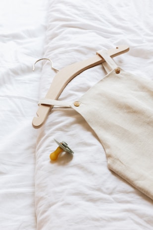 A wooden hanger holds a beige linen garment, laid out on a soft white surface, next to a yellow pacifier with a gray rim.